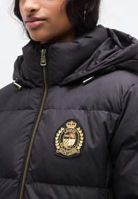 Μαύρο μπουφάν τύπου puffer με ψηλό γιακά, με χρυσό κεντημένο λογότυπο και φερμουάρ. Το ύφασμα φαίνεται λείο και μονωμένο.