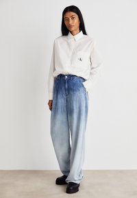 Λευκό πουκάμισο με γιακά και τσέπη, φέρει λογότυπο, συνδυασμένο με ψηλοκάβαλο μπλε τζην παντελόνι με κλίμακα χρωμάτων, και μαύρα παπούτσια με πλατφόρμα.