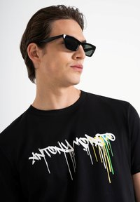 Camiseta negra con texto goteando en blanco y colores. El modelo lleva gafas de sol negras angulares, el cabello peinado hacia atrás, contra un fondo blanco.