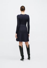 Πλεκτό φόρεμα σκούρο μπλε ναυτικό με μακριά μανίκια, υφή με σχέδιο, εφαρμοστή μέση, φαρδύς ποδόγυρος και στενή ζώνη. Συνδυασμένο με μαύρα μπότες μέχρι το γόνατο.
