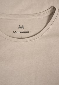 Beige pamut póló kerek nyakkivágással. Fekete nyomott "Matinique" logóval és "L" mérettel rendelkezik. Simább textúra, varrási részletekkel.