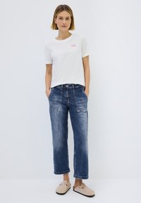 Weißes T-Shirt mit pinkem Text, blaue Jeanshose mit lockerer Passform und gestickten Taschen sowie beigen Clogs mit einer Schnalle.