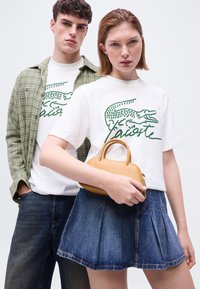 Νεαρή γυναίκα και άνδρας που φορούν λευκά γραφικά T-shirts Lacoste· η γυναίκα κρατά μικρή ταμπά τσάντα χειρός και φορά τζιν φούστα, ο άνδρας φορά πράσινο καρό πουκάμισο.