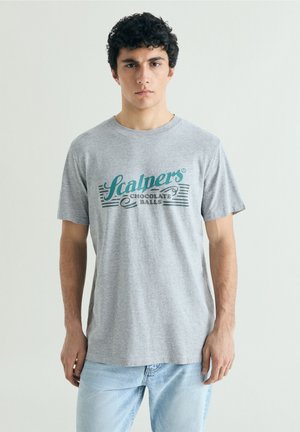 Jeune homme aux cheveux bouclés et foncés, portant un t-shirt gris avec un texte bleu sarcelle et noir et un jean bleu clair, debout devant un fond uni.