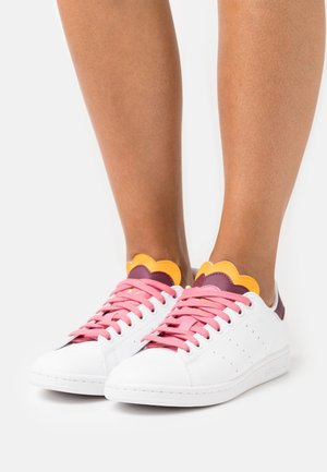 White sneakers with pink laces and scalloped yellow and purple tongue details worn on bare feet against a white background.