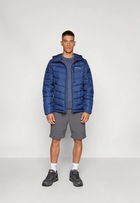 Navy blue puffer jacket with a zip front and hood, paired with a gray t-shirt and shorts. Model wears gray sneakers.
