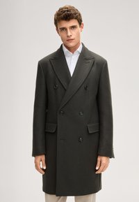Donkergroene dubbelgeknoopte jas met schoudervullingen, reverskraag met inkeping en zwarte knopen. Gladde wollen textuur en zichtbare zijzakken.