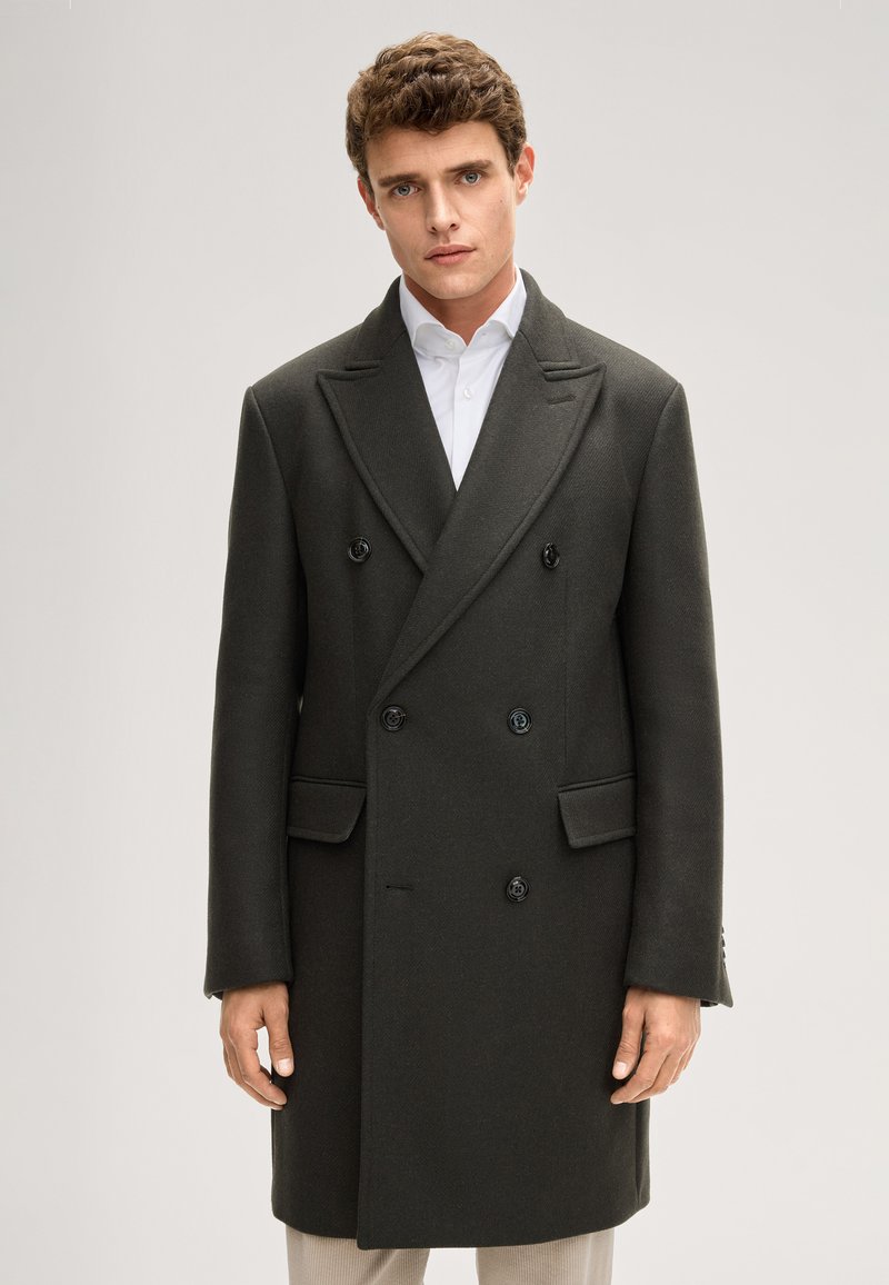 Donkergroene dubbelgeknoopte jas met schoudervullingen, reverskraag met inkeping en zwarte knopen. Gladde wollen textuur en zichtbare zijzakken.