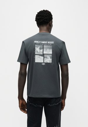 Férfi sötétszürke "World Famous Dickies" feliratú pólóban, melynek hátulján négy fekete-fehér vintage munkaképet nyomtak.