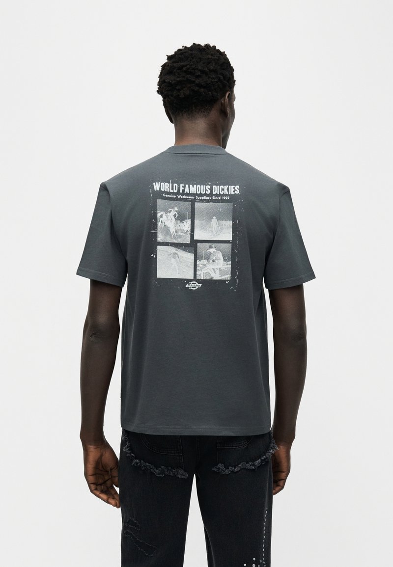 Hombre con camiseta de color gris oscuro "World Famous Dickies" con cuatro escenas vintage de trabajo en blanco y negro impresas en la parte posterior.