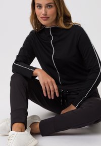 Black long-sleeve top with a high collar and silver piping along the front and sleeves, paired with black trousers and white sneakers.