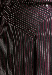 Tessuto a righe, principalmente nero con linee rosse e bianche. Presenta cuciture angolari e una texture liscia. Dettaglio di ferraglia visibile sulla cintura.