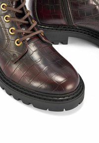 Brązowe botki za kostkę z ekoskóry z krokiem węża, złotymi oczkami i ciemnymi gumowymi podeszwami, z sznurowadłami i suwakiem po boku.