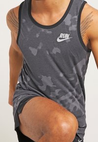 Un athlète portant un débardeur noir de Nike "RUN" et des shorts noirs, levant un genou comme s'il s'échauffait ou s'étirait.