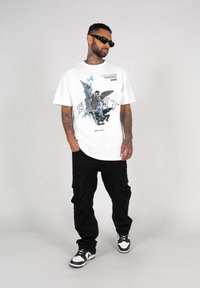 Hvid grafik t-shirt med et farverigt print af en figur med vinger, parret med sorte cargobukser og sort-hvide sneakers.