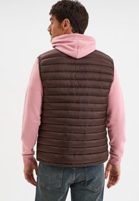 Braune Pufferweste mit horizontalem Steppmuster, kombiniert mit einem hellrosa Hoodie. Verfügt über einen hohen Kragen und keine sichtbaren Beschläge.
