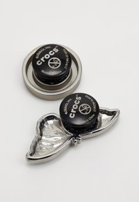 Due jibbets rotondi neri del marchio Crocs, uno in metallo lucido con un design curvo simile a un'ala e l'altro su una base circolare argentata.