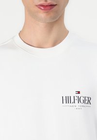 Hvit bomullssweatshirt med rund hals; har en svart "HILFIGER"-logo, ledsaget av røde og blå aksenter over teksten.