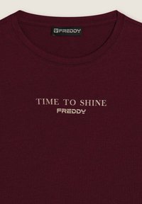 Camiseta de algodón burdeos con cuello redondo, con las inscripciones "TIME TO SHINE" y "FREDDY" en gris claro. Textura suave, diseño informal.