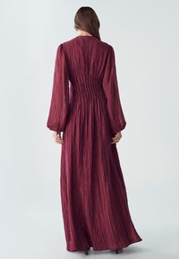 Dlouhý rukáv, vínová šaty s texturovanou tkaninou, mají přiléhavý korzet a splývavou sukni. Vertikální záhyby dodávají detail a pohyb.