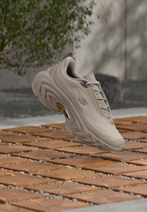 Beige athletic sneaker with textured sole suspended above wooden tiles outdoors, with blurred wall and plant in background.