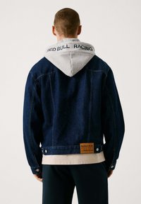 Chaqueta de denim en azul oscuro con capucha gris, que presenta un logo en la capucha y un parche de la marca en la parte trasera. Corte estándar con cierre de botón.