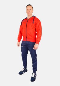 Sportowy dres składający się z jaskrawej czerwonej bluzy z zamkiem błyskawicznym i granatowymi akcentami oraz pasujących granatowych spodni dresowych. Noszony z czarnymi butami sportowymi.