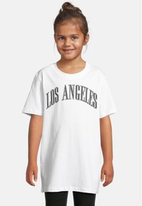 Balta medvilninė marškinė su užrašu "LOS ANGELES" ryškiomis juodomis raidėmis, trumpomis rankovėmis ir laisvu siluetu, dėvima su juodomis leggingais.