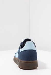 Zapatillas de ante marino con detalles en azul claro, suela de goma texturizada y tres rayas blancas en el lateral. La vista trasera resalta el diseño del talón.