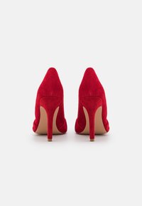 Röda mocka högklackade skor med spetsig tå och en slank klack. Texturen är mjuk, med en len insida och en beiget sula.