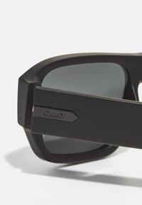 Gafas de sol rectangulares negras con lentes oscuros, con un marco texturizado. El lado izquierdo muestra un destacado logo "QUAY".