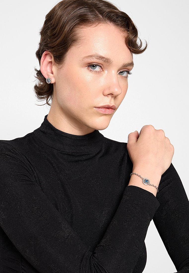 Srebrna bransoletka z okrągłym niebieskim kamieniem szlachetnym, noszona na nadgarstku, obok kolczyków wkręcanych z podobnymi niebieskimi kamieniami. Czarny, strukturalny golf.