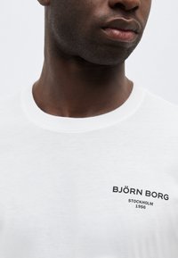 Κοντινό πλάνο άνδρα που φοράει λευκό πουκάμισο με την επιγραφή "BJÖRN BORG STOCKHOLM 1956" κοντά στο γιακά στην αριστερή πλευρά.