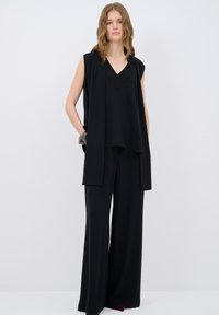 Zwarte mouwloze top met een V-hals en gelaagd ontwerp, gecombineerd met een wijdere zwarte broek. Glad materiaal met subtiele details op de top.