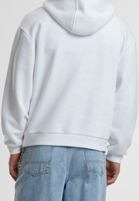 Felpa bianca in cotone con tasca a marsupio, polsini e orlo a costine. Indossata con pantaloni in denim di un blu chiaro. La vista da dietro mostra i dettagli del cappuccio.