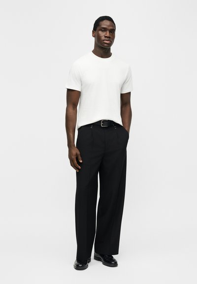 Homme debout portant un t-shirt blanc uni à manches courtes, une ceinture noire, un pantalon noir à jambes larges, et des chaussures habillées noires, les mains dans les poches.