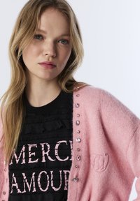 Jasnoróżowy sweter dzianinowy z ozdobnymi koralikami przy zapięciu na guziki, noszony na czarnej koszuli o fakturze z różowym napisem.