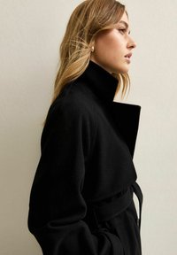 Abrigo negro de lana con cuello alto, cintura con cinturón y detalles de botones. La tela tiene una textura suave, creando una silueta elegante.