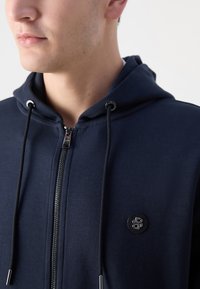 Marineblauwe zip-up hoodie met een trekkoordcapuchon en zilverkleurige hardware. Bevat een logopatch op de linkerborst en een gladde katoenmix textuur.
