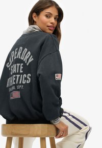 Felpa nera con stampa "SUPERDRY STATE ATHLETICS" in grigio, toppe con bandiera degli Stati Uniti e polsini a coste. Modello seduto su uno sgabello di legno.