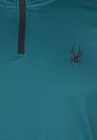 Sudadera verde azulado con un cierre negro y un pequeño logo de araña negra en el pecho. La tela tiene un patrón texturizado y transpirable.