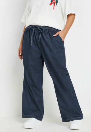 Person trägt lockere dunkelblaue Jeanshose mit Kordelzug in der Taille, weiße Turnschuhe und ein weißes Oberteil mit kleinem roten und grünen Design.