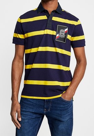 Muž v tmavě modré polotriko s vodorovnými žlutými pruhy a černým límcem, v kombinaci s modrými džíny, rukou v kapse.