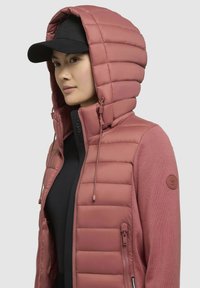Gevoerde roze jas met een gestructureerde capuchon, rits aan de voorkant en een rond logo-opdruk op de mouw. Een zwarte pet maakt de outfit compleet.
