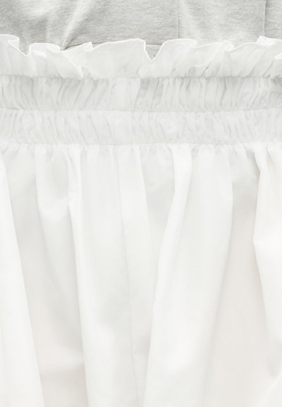 Λευκό ύφασμα με ελαστική ζώνη μέσης και σούρες, φορεμένο πάνω από ανοιχτό γκρι ένδυμα.