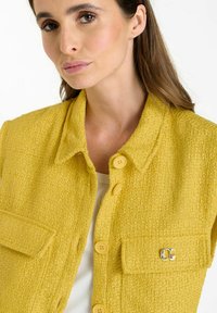 Veste texturée jaune avec un col, dotée de boutons jaunes et d'un accent argenté sur la poche. Fabriquée en tissu tissé.
