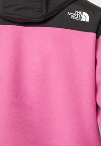 Chaqueta de forro polar con un cuerpo rosa y sección superior negra, con capucha. Logo blanco en el hombro izquierdo, textura suave.