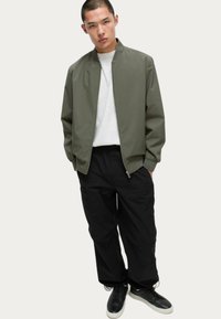 Chaqueta bomber verde oliva con cuello ajustado, cierre de cremallera y puños elásticos, llevada sobre una camisa blanca y combinada con pantalones cargo negros y zapatillas deportivas.