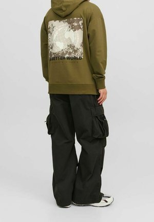 Person iført olivengrøn hættetrøje med jordklode og teksten "A Better World" på ryggen, sorte cargobukser og hvide sneakers, stående sidelæns.