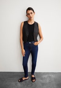 Un gilet sans manches noir avec des boutons en bois, associé à un jean taille haute bleu foncé et des sandales noires à enfiler, se tenant devant un fond neutre.